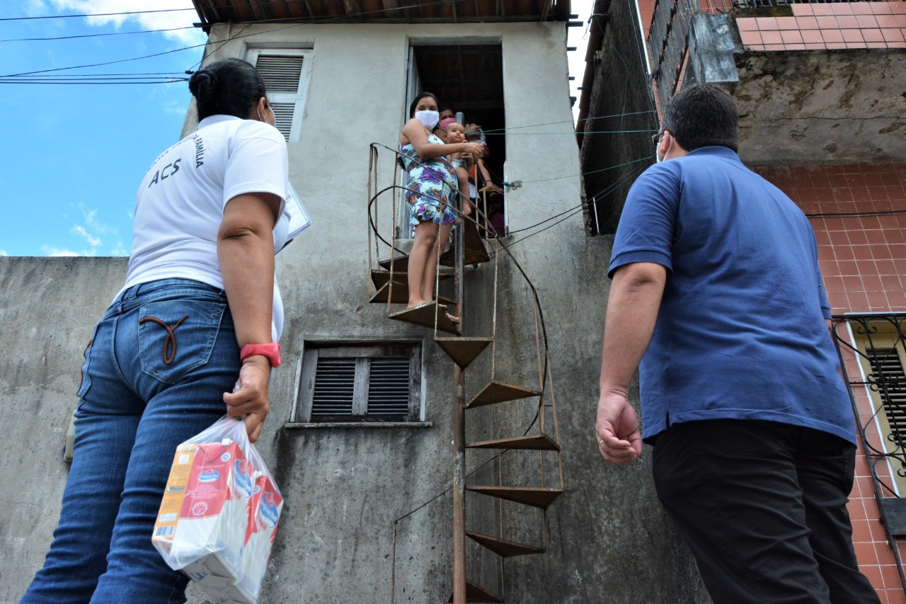 Duas pessoas com máscara na porta de uma casa onde se encontra uma mulher com uma bebê no colo